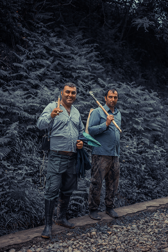Two railway workers walking along the tracks in northern Iran, carrying axes and smiling as the pass through forested mountains.