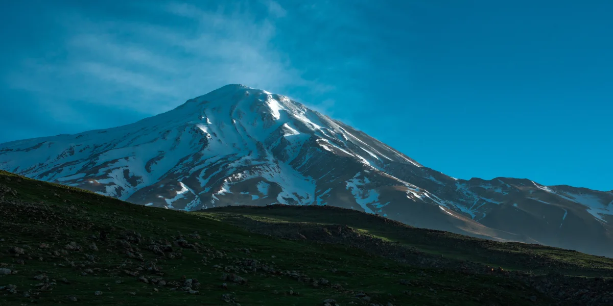 Mount Damavand at sunrise with a deep blue sky, soft clouds, and green pastures in the foreground - Iran