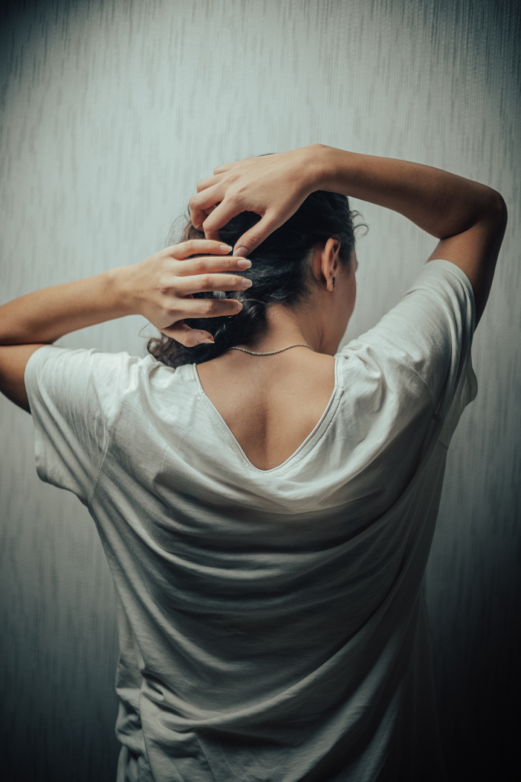 A young Iranian woman standing with her back to the camera, gathering her hair, she wears a loose, wrinkled white shirt that exposes part of her neck and upper back. a delicate necklace rests on her skin.