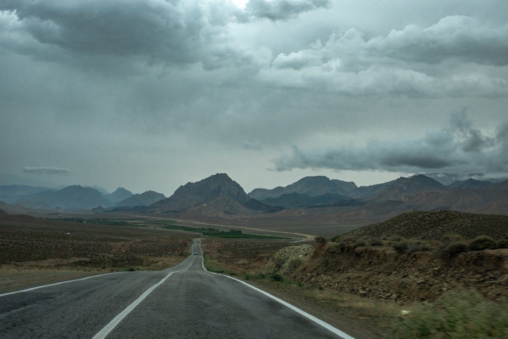 A long, straight road seen from above, stretching toward rocky mountains on the Damghan-Kiasar route.