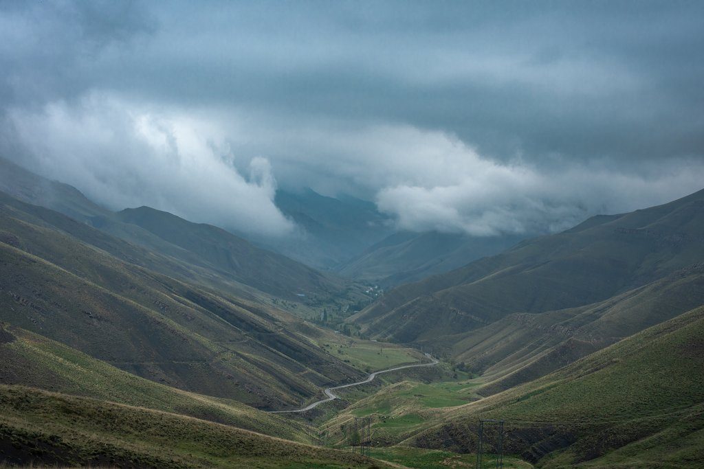 A wide aerial shot of Nesn village nestled deep in an Alborz valley, with clouds moving into the gorge and a road winding down from the mountain pass.