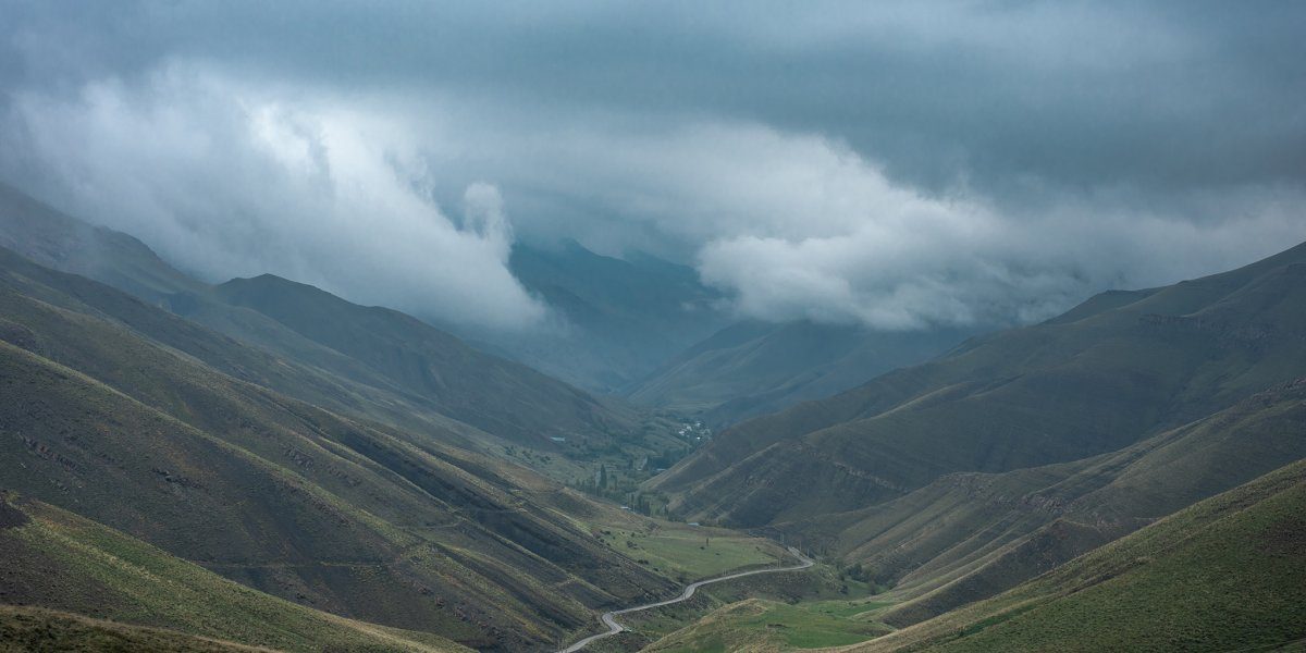 A wide aerial shot of Nesn village nestled deep in an Alborz valley, with clouds moving into the gorge and a road winding down from the mountain pass.