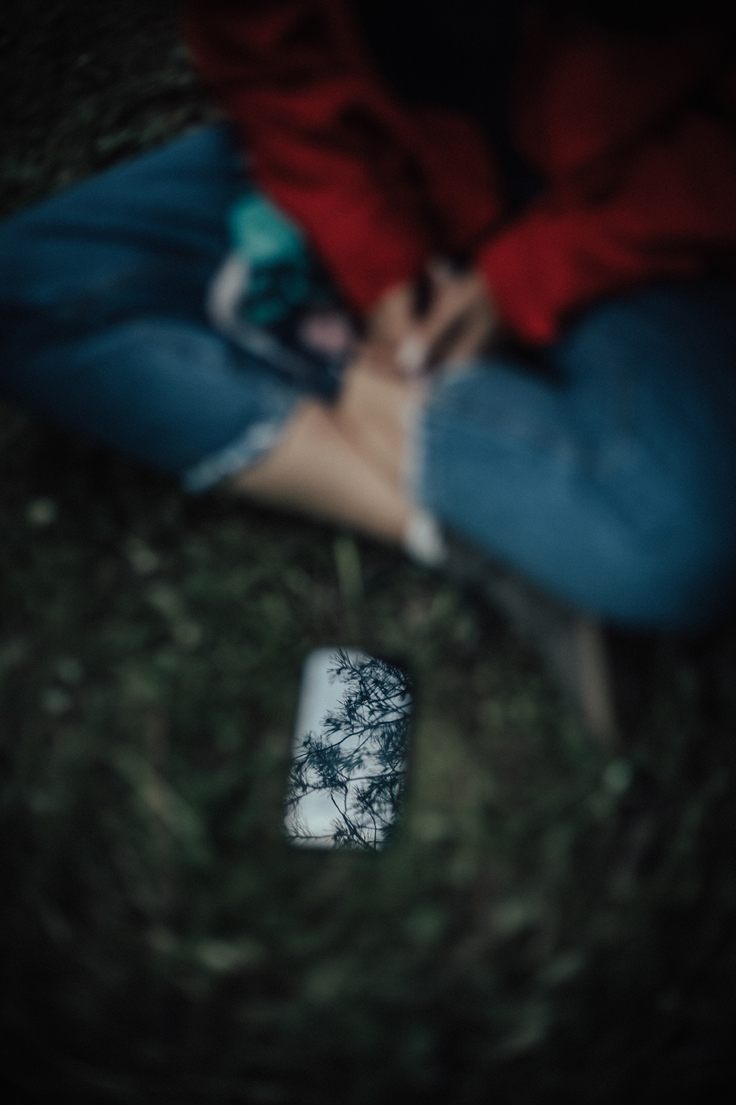 A girl in a red coat and jeans sitting on the ground with a smartphone reflecting tree branches, captured in shallow focus.