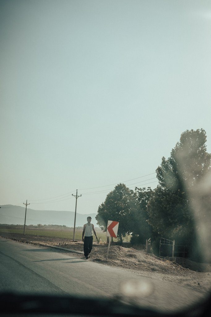 An elderly man walking along a rural road to Isfahan, next to road signs and electric tower replacing trees.